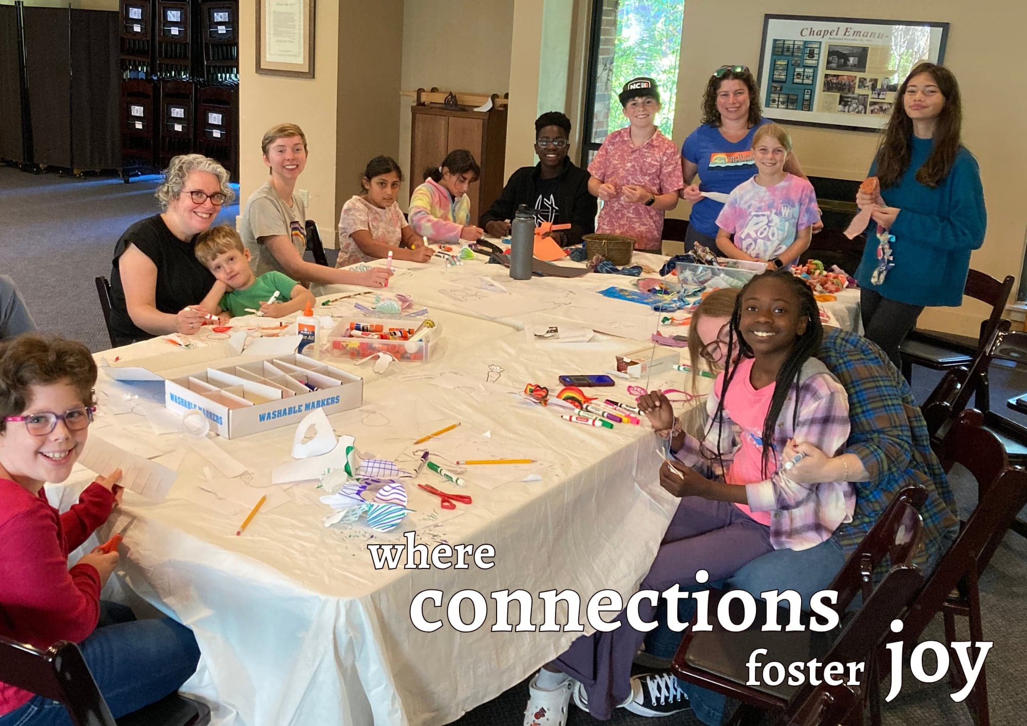 Children and adults making crafts together around a long table, with the words “where connections foster joy”