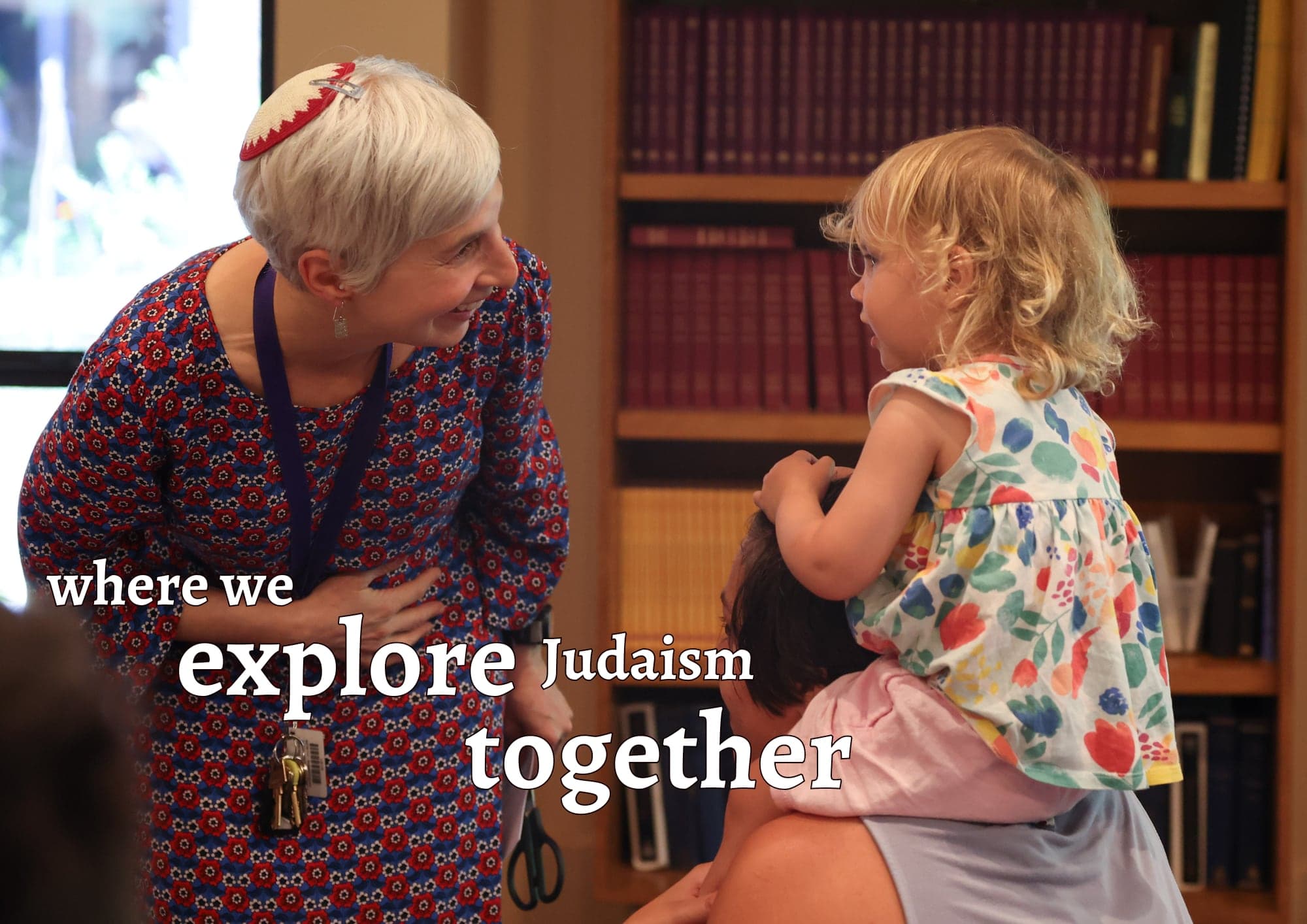 An adult in a kippah bending down to greet a toddler, with the words “where we explore Judaism together”