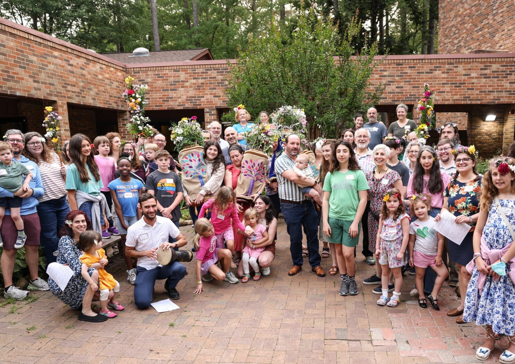 A large group of congregants of all ages gathered in a brick courtyard with flower-decorated Torah scrolls